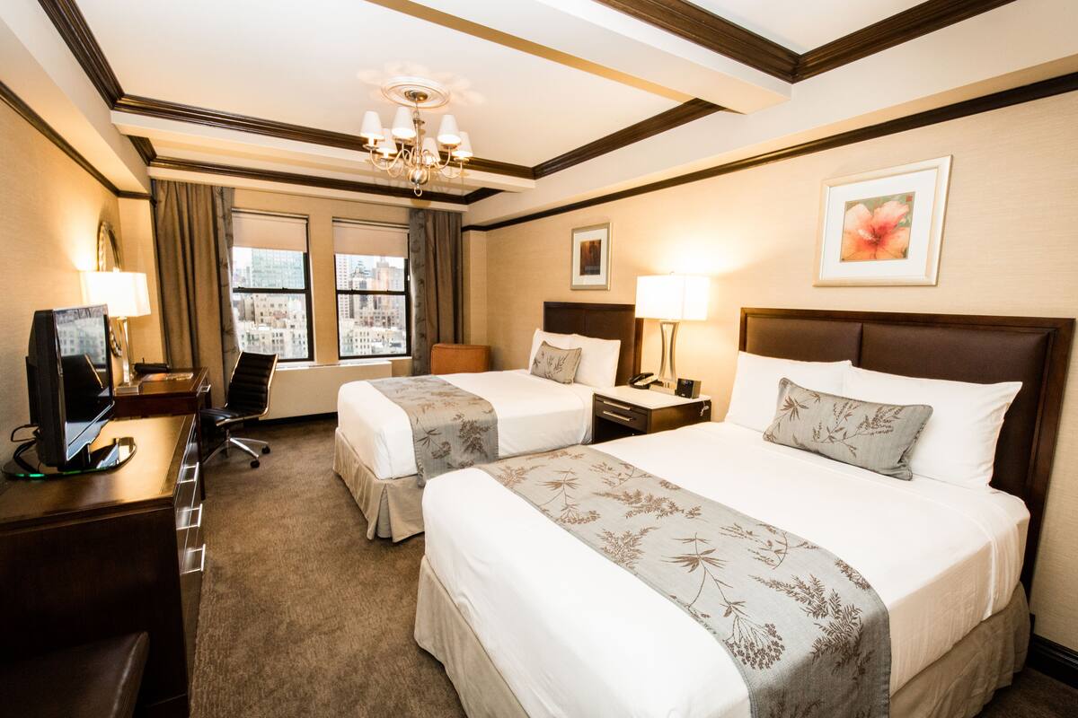 Quarto de hotel com duas camas, uma escrivaninha com computador, uma cadeira e janela com vista para a cidade.