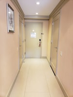 Een gang met beige wanden en een betegelde vloer, die leidt naar een deur met een rookmelder en een bord met een evacuatieplan.