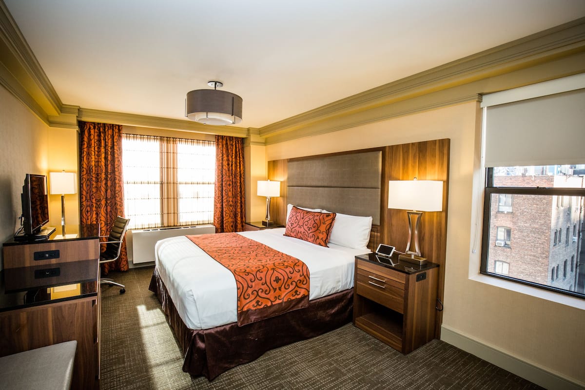 Quarto de hotel com uma cama grande, duas mesas de cabeceira, uma escrivaninha, uma cadeira, uma televisão e uma janela com vista para edifícios.