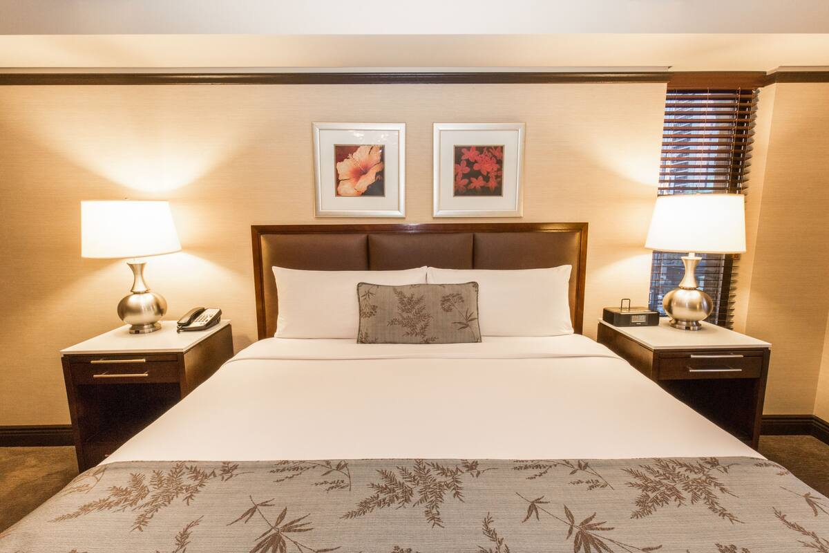 Quarto de hotel com uma cama grande, duas mesas de cabeceira com abajures, um telefone e duas quadros na parede.