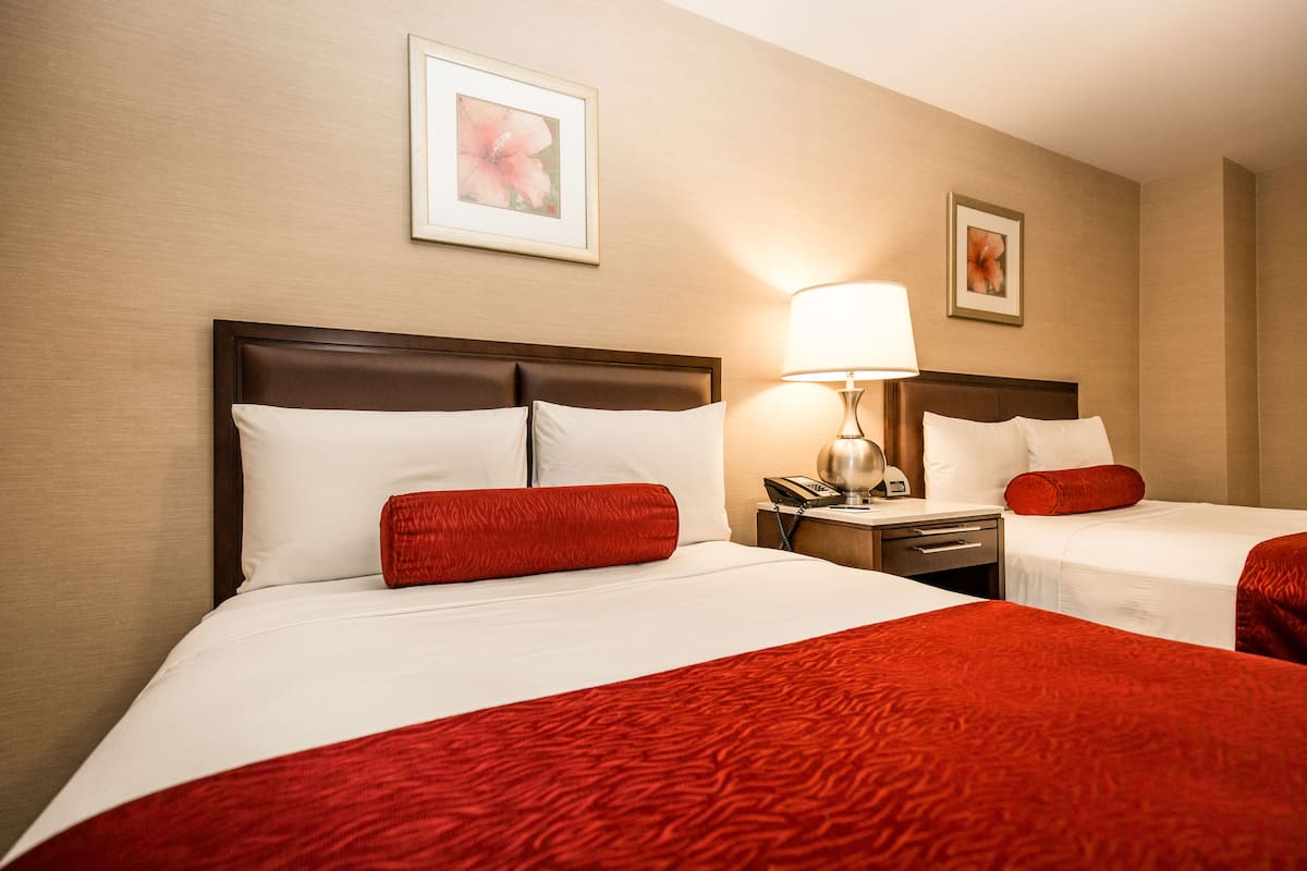 Quarto de hotel com duas camas, cada uma com roupa de cama branca e detalhes vermelhos, mesa de cabeceira com abajur e quadro com tema floral na parede.