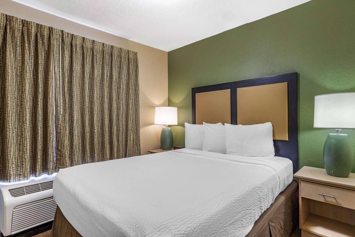 Habitación de hotel con una cama grande, dos mesitas de noche, una lámpara y una ventana con cortinas.