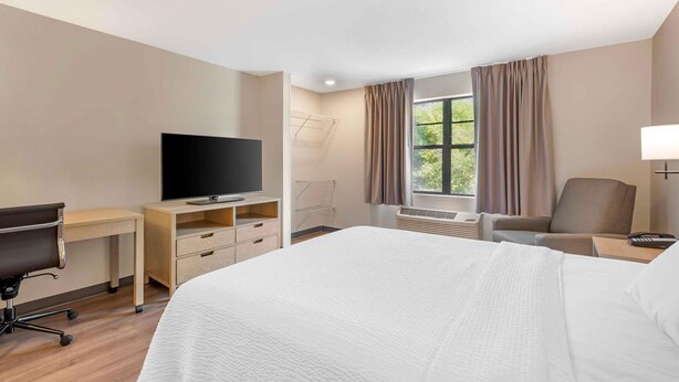 A hotel room with a bed, a desk with a TV, a chair, a window with curtains, and an air conditioning unit. - Extended Stay America Suites St Petersburg Clearwater ExecDr (Clearwater)