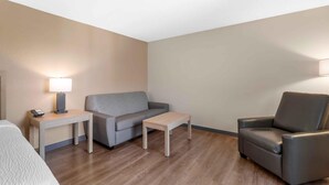 A hotel room with a grey sofa, a small table, a chair, and a lamp on a nightstand. - Extended Stay America Suites St Petersburg Clearwater ExecDr (Clearwater)