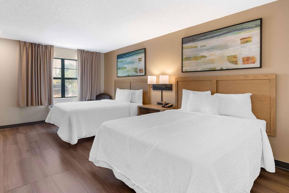 Habitación de hotel con dos camas, cabecero de madera, mesita de noche con lámpara, ventana con cortinas y dos cuadros colgados en la pared.