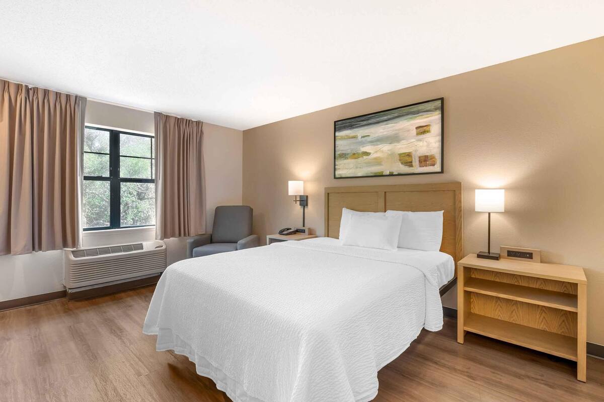 Habitación de hotel con cama, una silla, escritorio, lámpara y un cuadro en la pared.