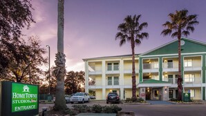 A multi-story hotel with a green and white facade, a signboard for HomeTown Studios, and a parking area with cars. - HomeTowne Studios by Red Roof Houston - Northwest (Houston)