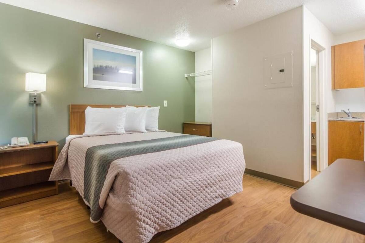 Ein Hotelzimmer mit Bett, Nachttisch, Lampe und Blick auf die Küche.