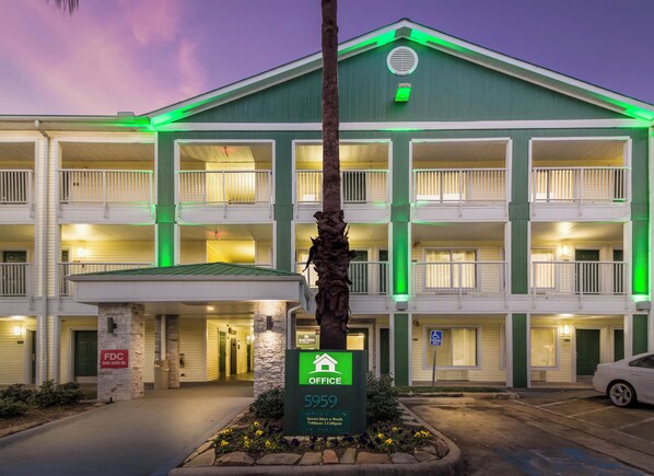 Un hotel de varios pisos con fachada verde, entrada central y señal de estacionamiento que indica tarifa semanal.