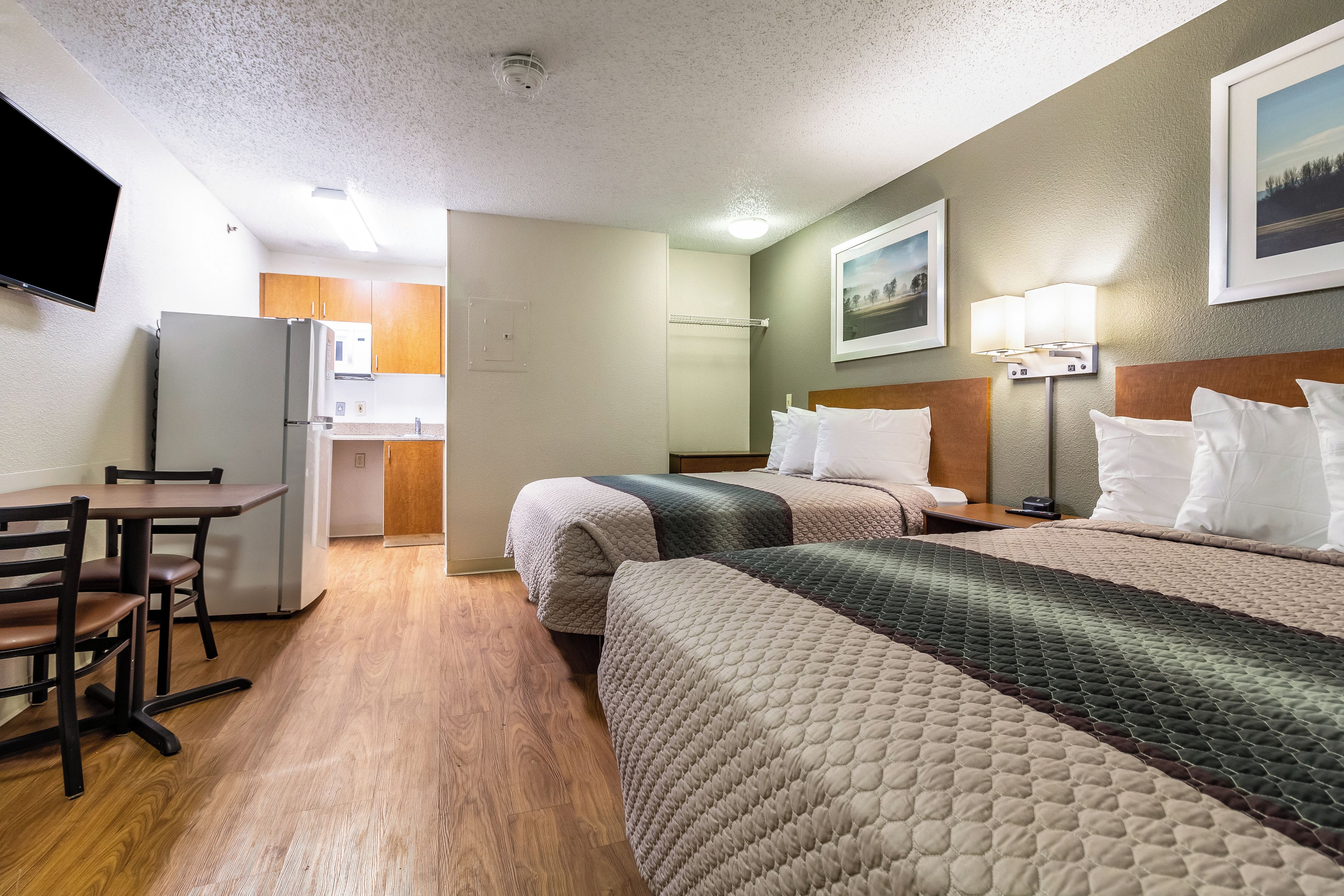 A hotel room with a kitchenette, a bed with multiple pillows, a desk with a chair, and a wall-mounted TV.