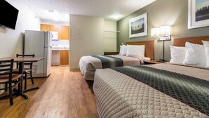 A hotel room with a kitchenette, a bed with multiple pillows, a desk with a chair, and a wall-mounted TV. - HomeTowne Studios by Red Roof Houston - Northwest (Houston)