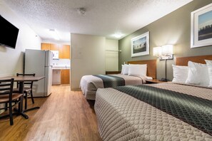 Habitación de hotel con kitchenette, cama con varias almohadas, escritorio con silla y televisor montado en la pared.
