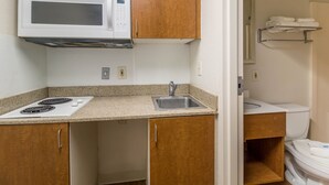 A compact kitchen with a microwave, stove, sink, and wooden cabinets. - HomeTowne Studios by Red Roof Houston - Northwest (Houston)