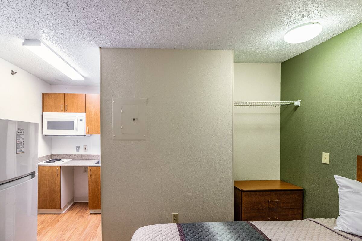 Une chambre d’hôtel compacte, dotée d’une kitchenette, d’un lit avec une couverture matelassée et d’un mur d’accent de couleur verte.