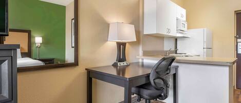 A hotel room with a desk, chair, lamp, kitchenette, and a large mirror.