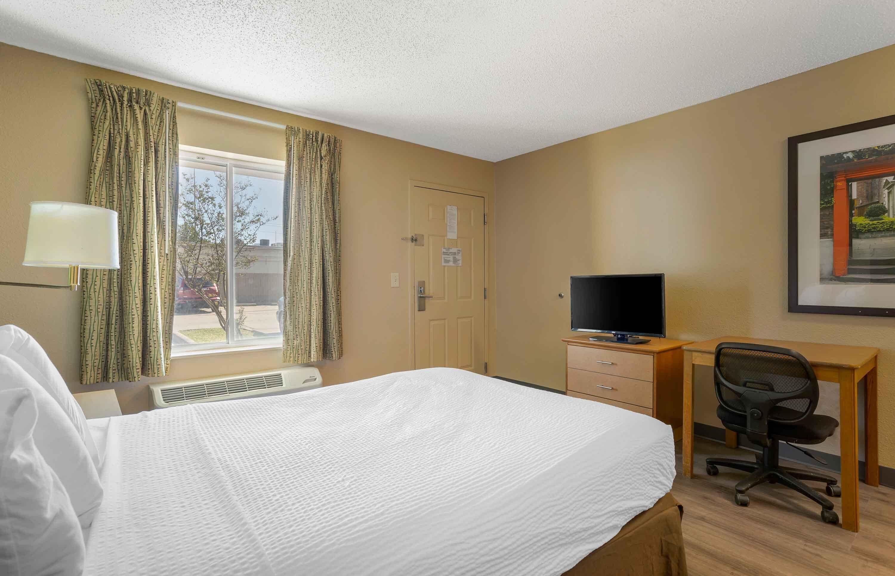 A hotel room with a bed, a desk with a chair, a television, and a window with curtains.