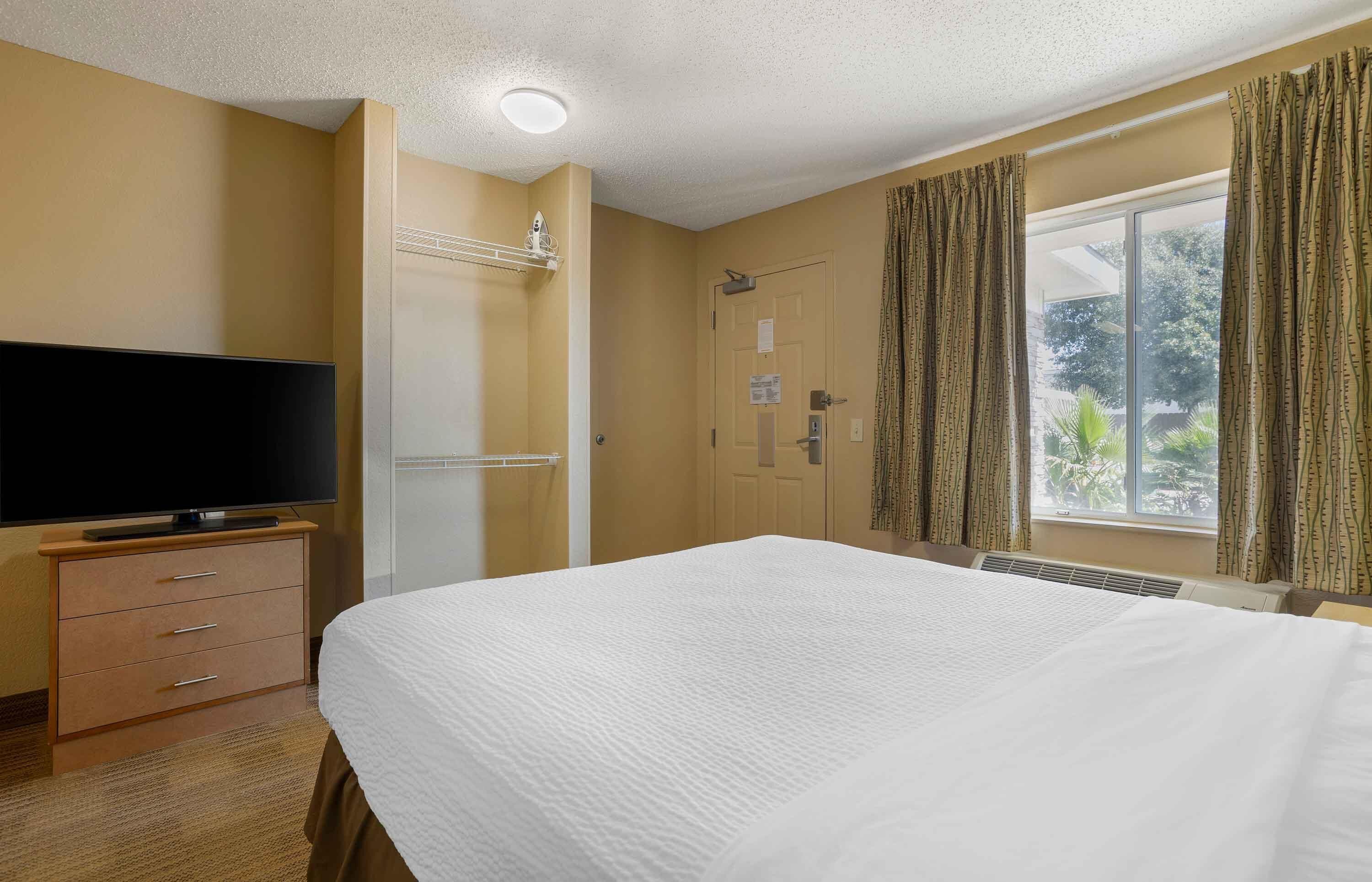 Una habitación de hotel con una cama, un televisor sobre un mueble de madera, una puerta con toallero y una ventana con cortinas.