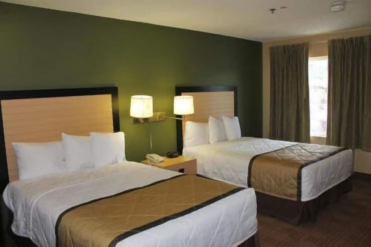 A hotel room with two beds, a nightstand with a lamp, and a window with curtains.