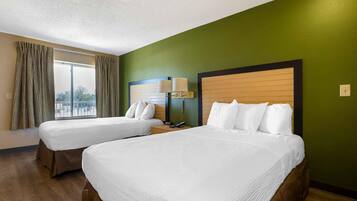 A hotel room with two beds, a window with curtains, a nightstand with a lamp, and a view of the outdoors.