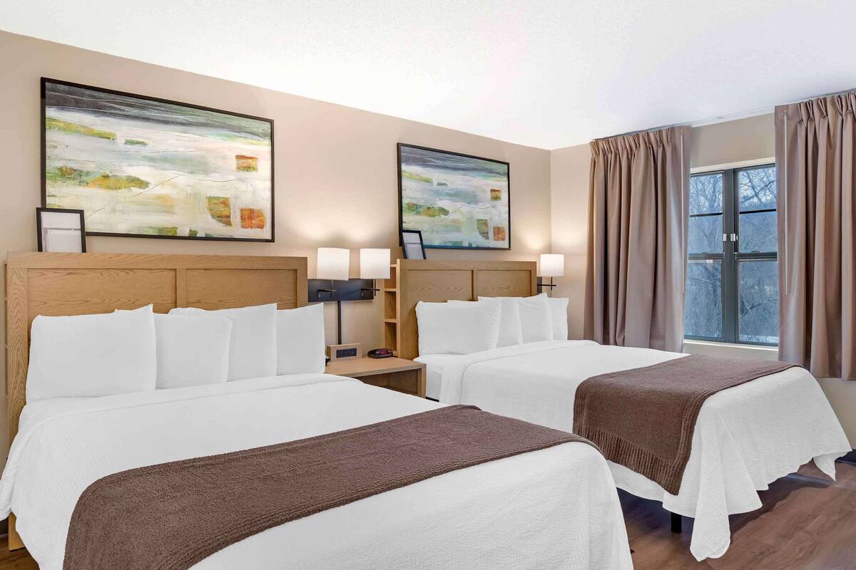 Une chambre d’hôtel avec deux lits, une tête de lit en bois, une table de chevet avec une lampe et une fenêtre avec des rideaux.