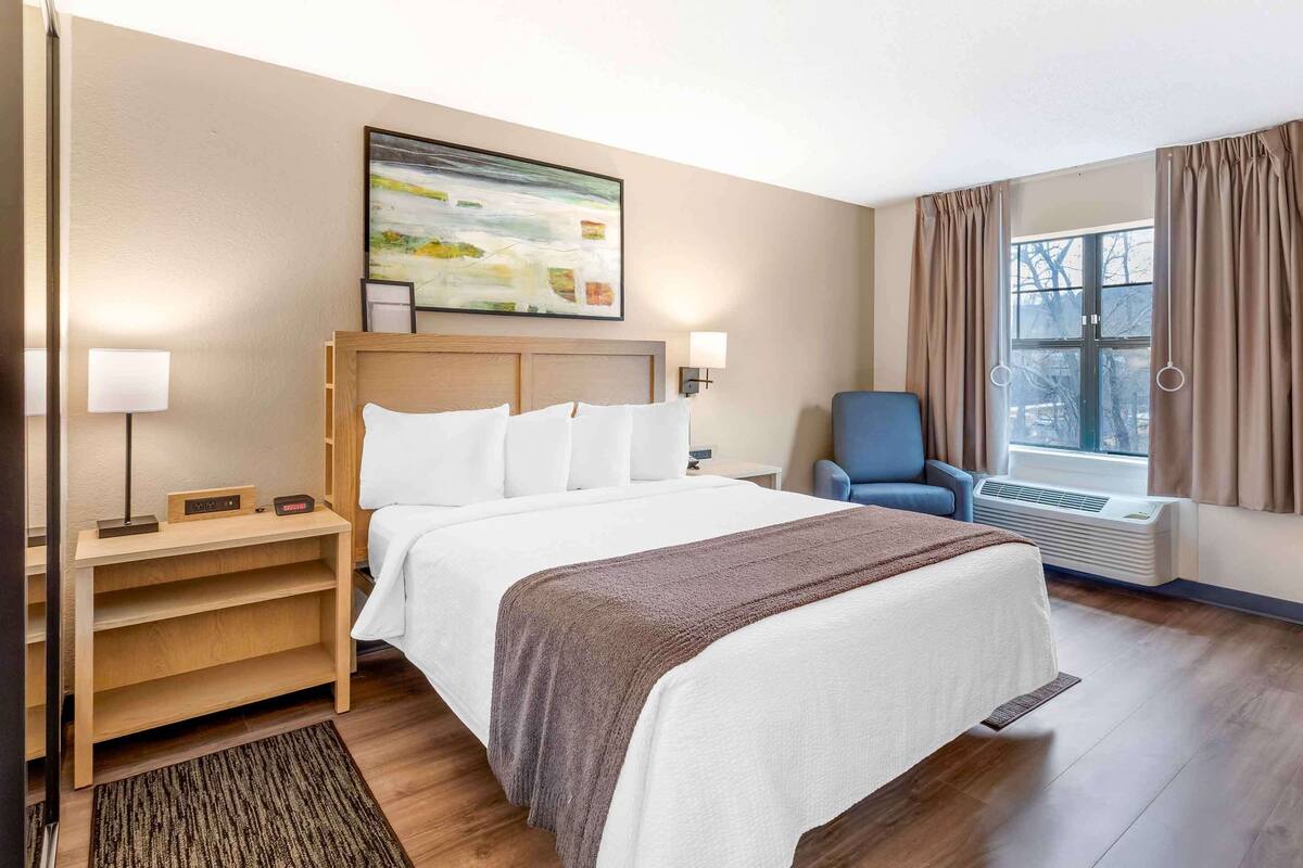 Une chambre d’hôtel avec un lit, une chaise, un bureau avec une lampe, un tableau au mur et une fenêtre avec des rideaux.