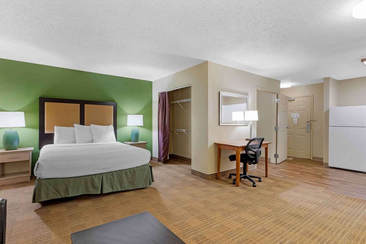 A hotel room with a bed, desk, chair, and a refrigerator.