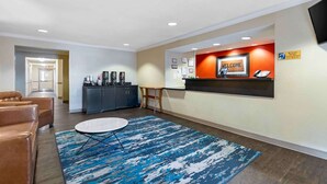 Un hall d’hôtel comprenant un comptoir d’accueil, un espace où l’on peut se servir du café, un coin salon avec un canapé et une table basse, ainsi qu’un mur d’accueil orné de tableaux et d’une pancarte de bienvenue.
