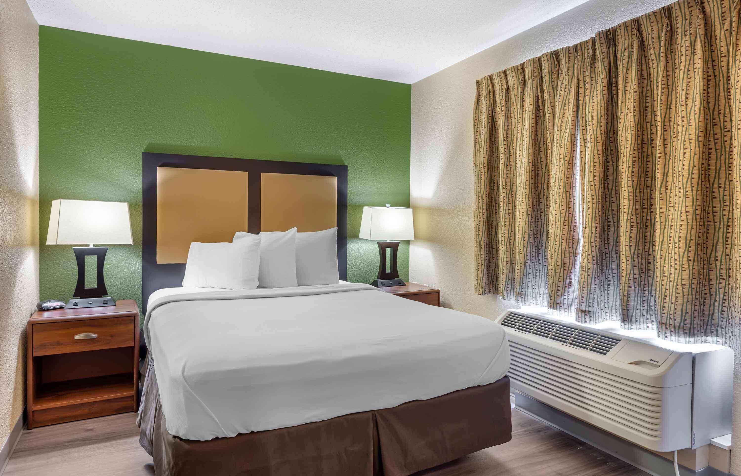 Habitación de hotel con una cama grande, dos mesitas de noche con lámparas, aire acondicionado de pared y una cortina estampada.