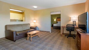 A hotel room with a sofa, a small table, a desk with a chair, a television, and a lamp. - Suburban Studios Macon North (Macon)