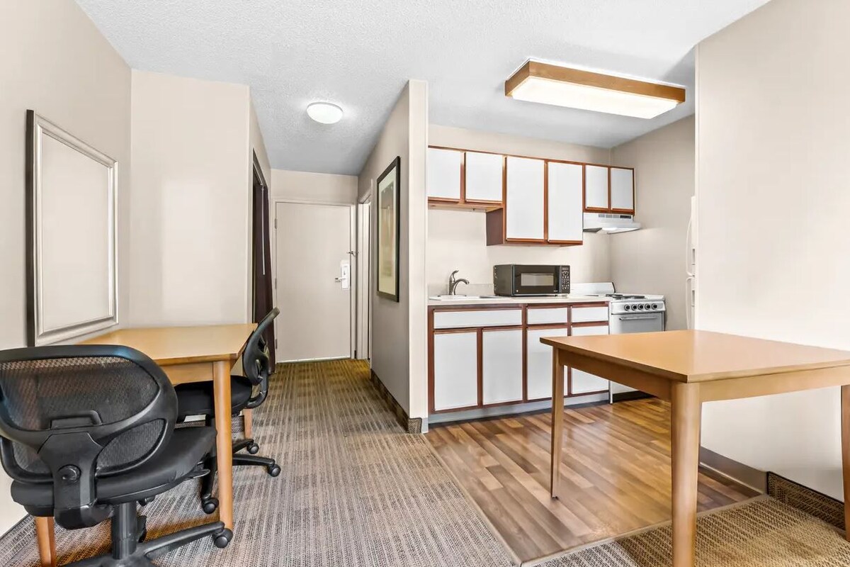 A compact room with a kitchenette, a desk with a chair, and a wooden floor.