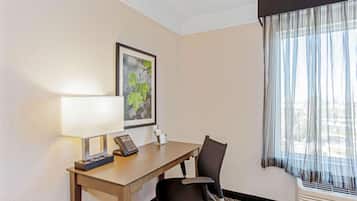 A hotel room with a desk, chair, lamp, framed picture, and a window with sheer curtains.