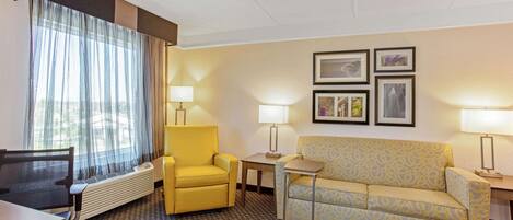 Una habitación de hotel con un sillón amarillo, un sofá estampado, un escritorio y una ventana con cortinas translúcidas.