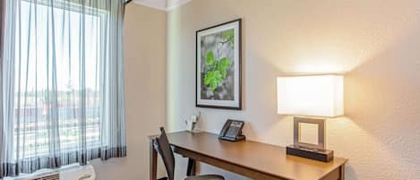 Habitación de hotel con escritorio, silla, aire acondicionado, ventana con cortinas traslúcidas, un cuadro en la pared y una lámpara de noche.