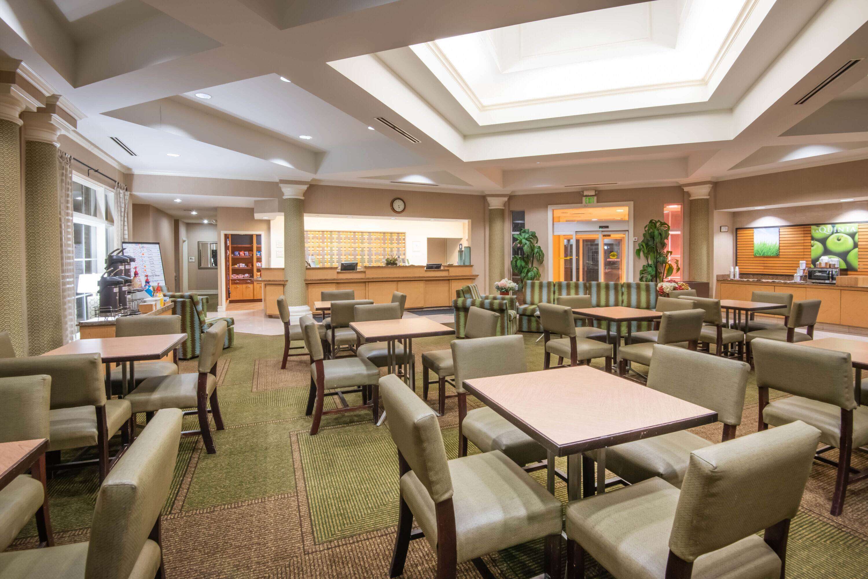 A hotel lobby with multiple seating areas, a reception desk, and a visible kitchen area.