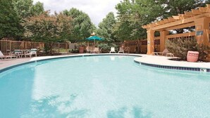 Piscina cristalina com espreguiçadeiras e guarda-sóis ao redor, cercada por árvores e uma pérgola de madeira.