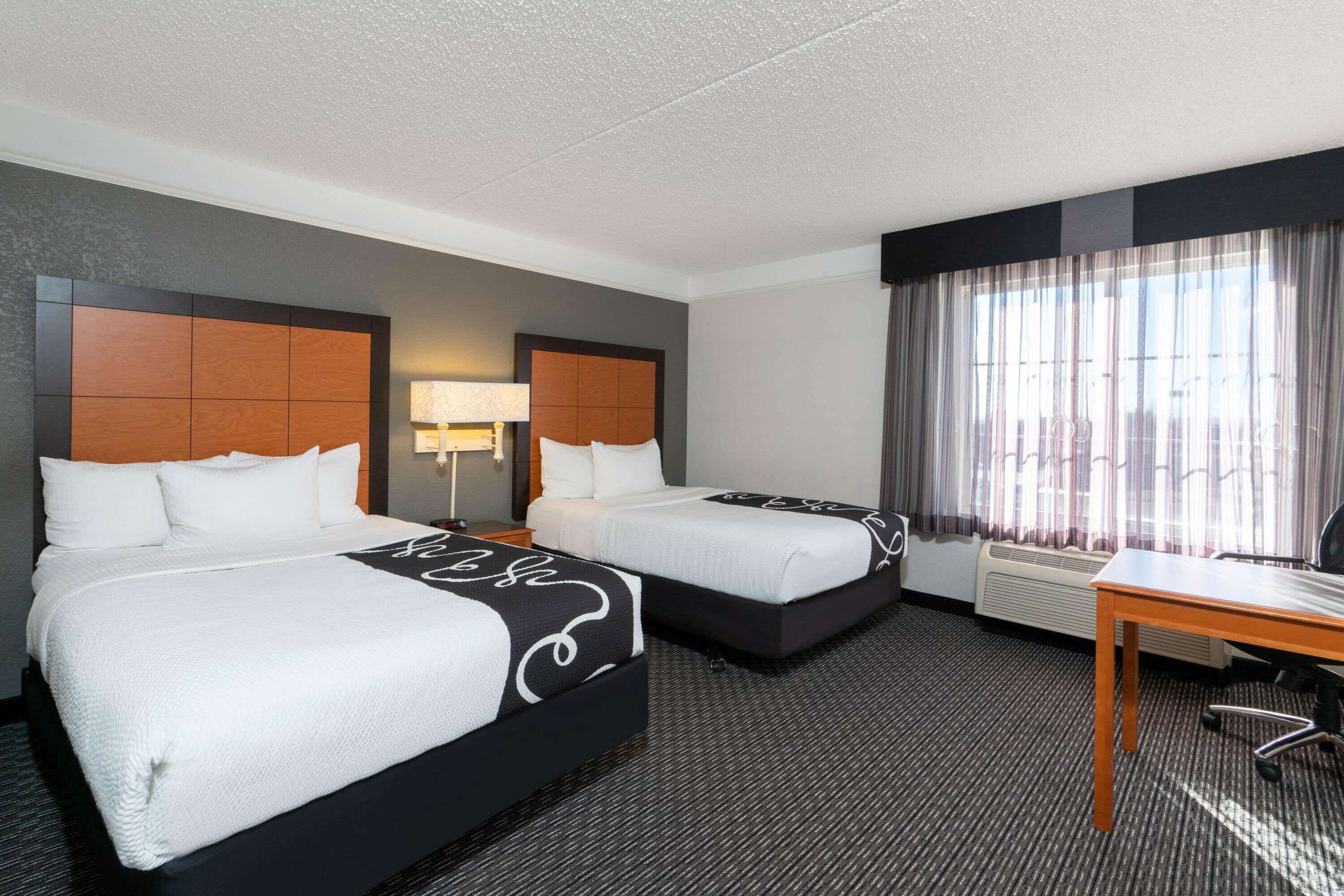 A hotel room with two beds, a desk, and a large window with curtains.