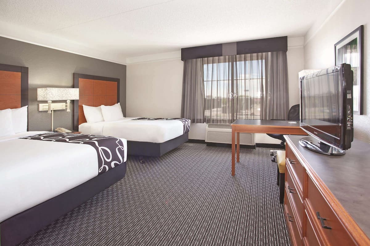A hotel room with two beds, a desk with a television, a lamp, and a large window with curtains.