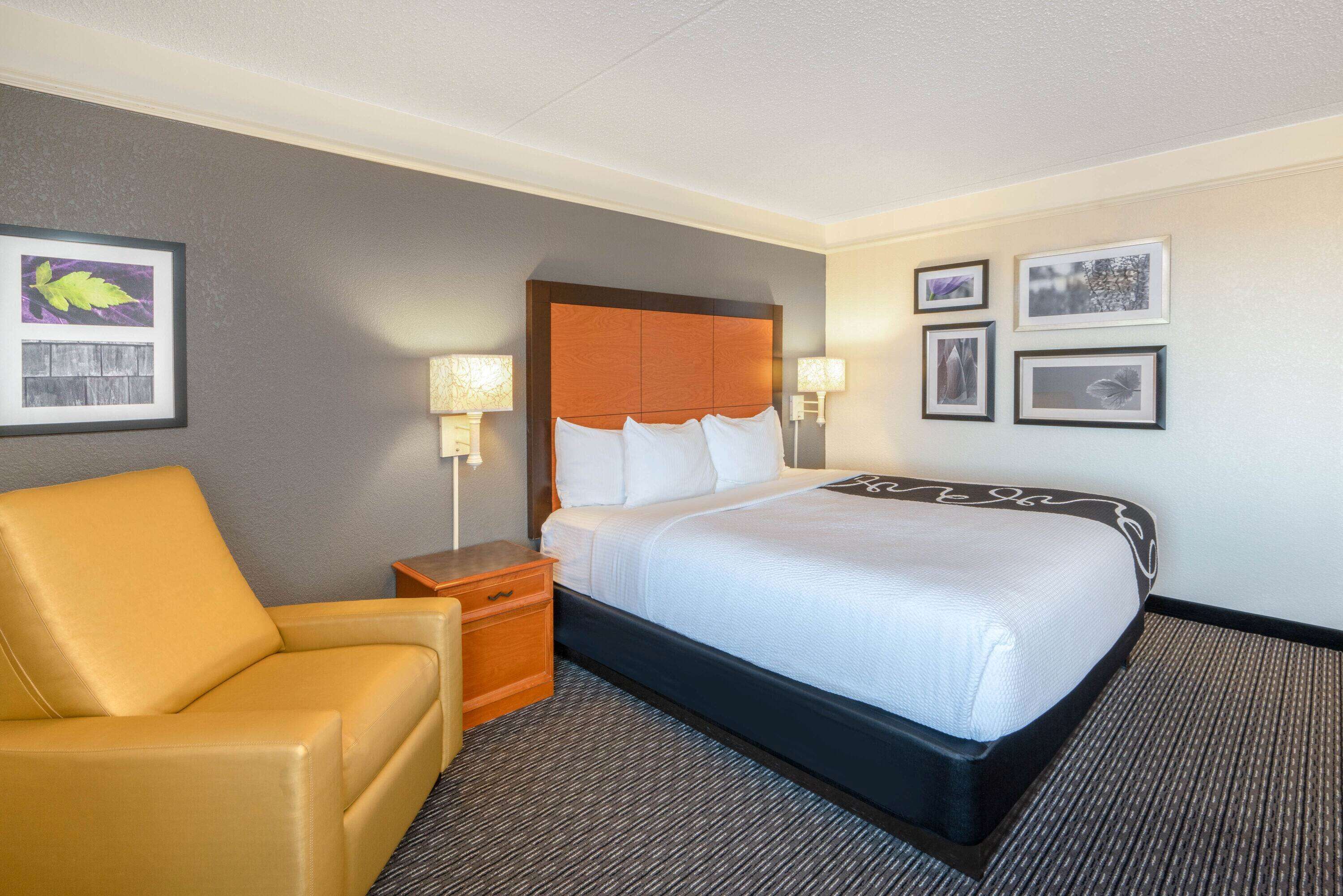 A hotel room with a bed, a chair, a nightstand, and framed pictures on the wall.