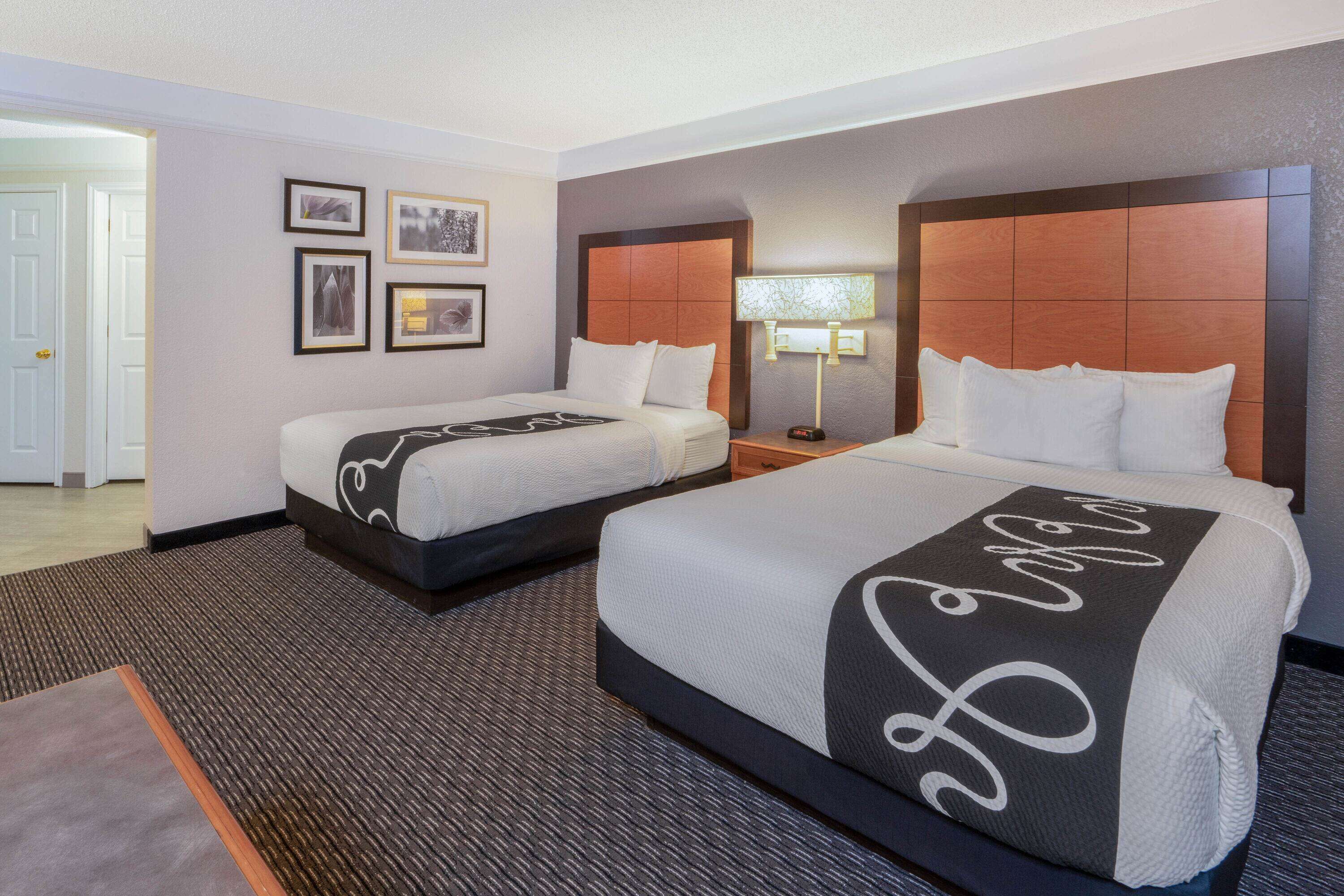 Habitación de hotel con dos camas, una grande con cabecera decorativa, una más pequeña, una mesita de noche con lámpara y cuadros en la pared.