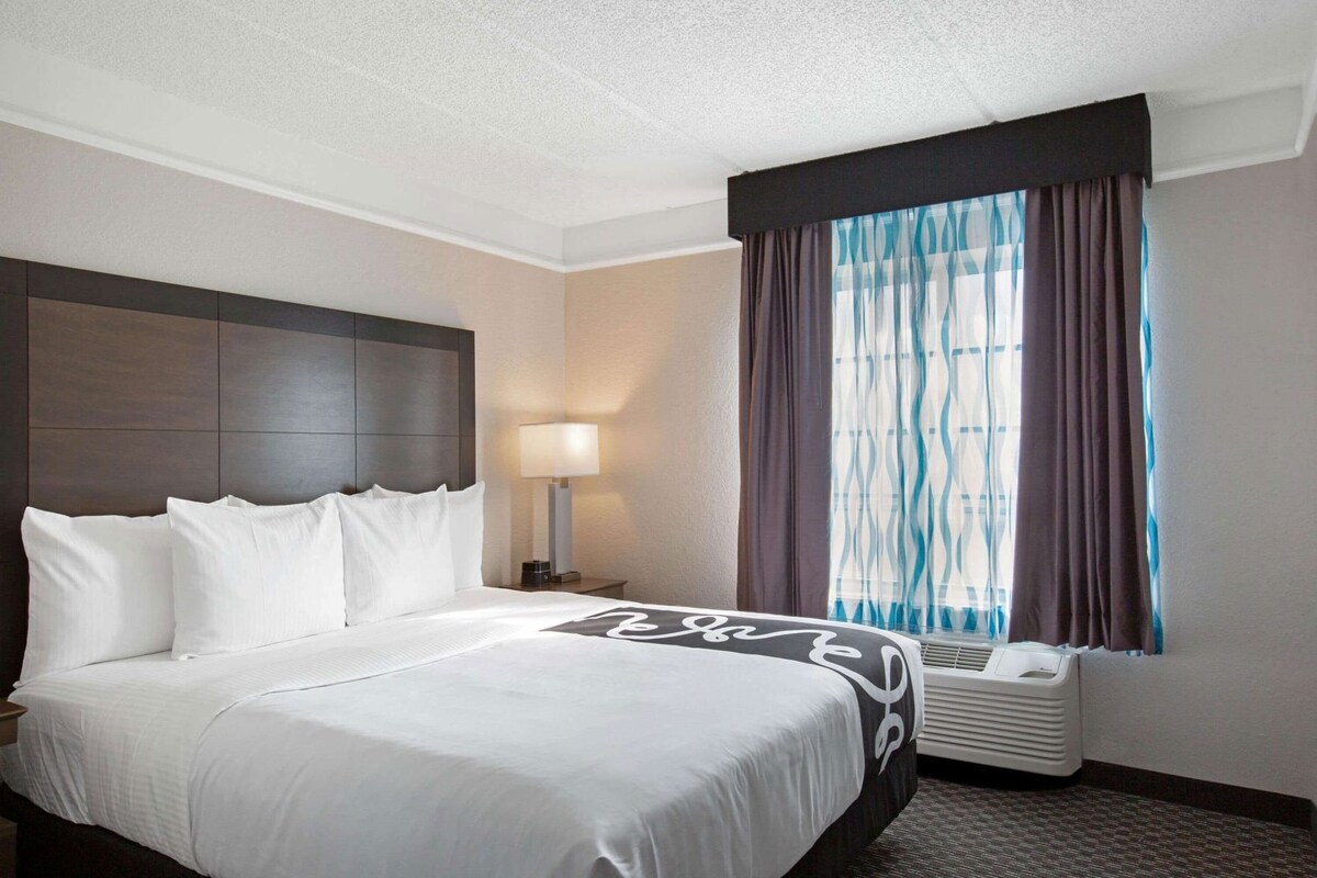 Une chambre d’hôtel avec un grand lit, une table de chevet avec une lampe, une fenêtre avec des rideaux et un thermostat fixé au mur.