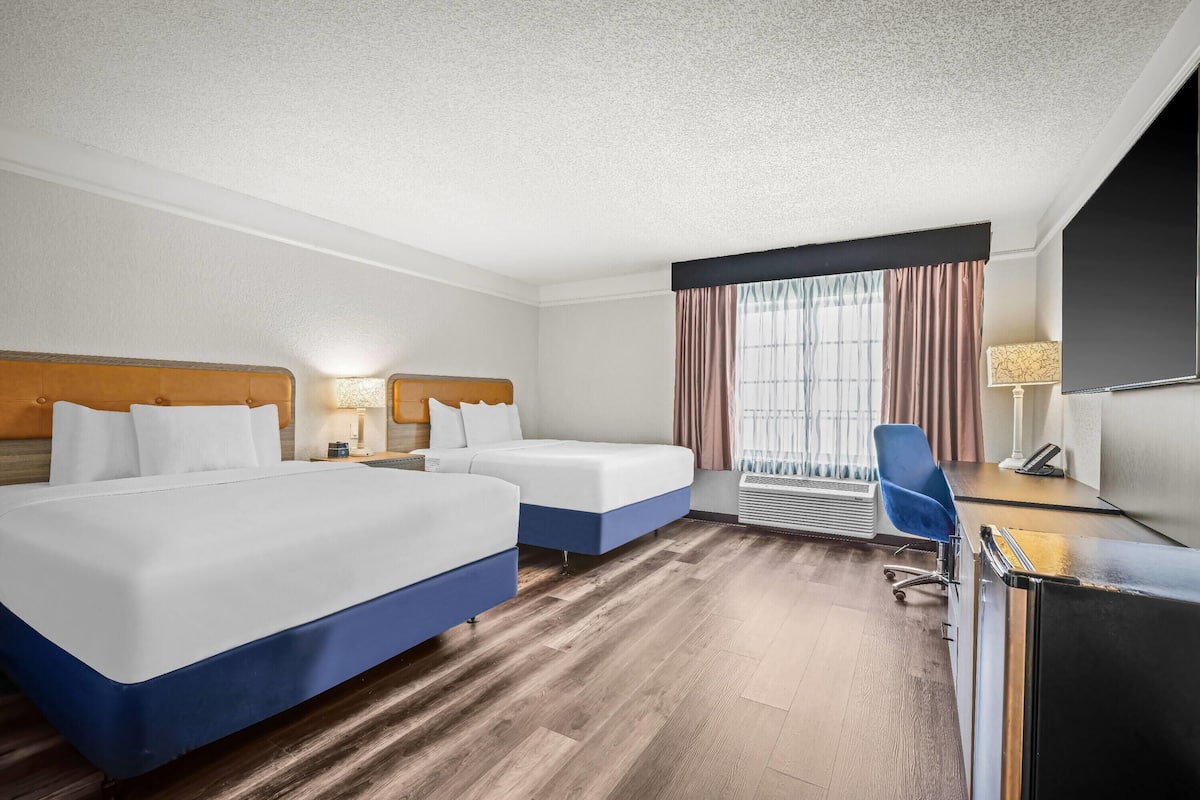 Ein Hotelzimmer mit zwei Betten, einem Schreibtisch, einem Stuhl, einem Fernseher und einem Fenster mit Vorhängen.