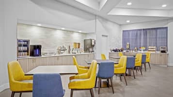 A modern hotel dining area with yellow and blue chairs, a long counter with a coffee machine, and a view of the kitchen in the background.