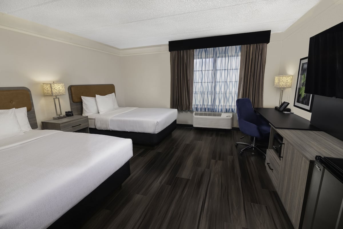 Ein Hotelzimmer mit zwei Betten, einem Schreibtisch, einem Stuhl, einem Fernseher und einem Vorhangfenster.