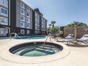 Zona de piscina de hotel con hidromasaje, sillas de descanso y un edificio de varios pisos y ventanas.