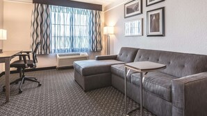 A hotel room with a sectional sofa, a small table, a desk with a chair, a standing lamp, and a large window with patterned curtains.
