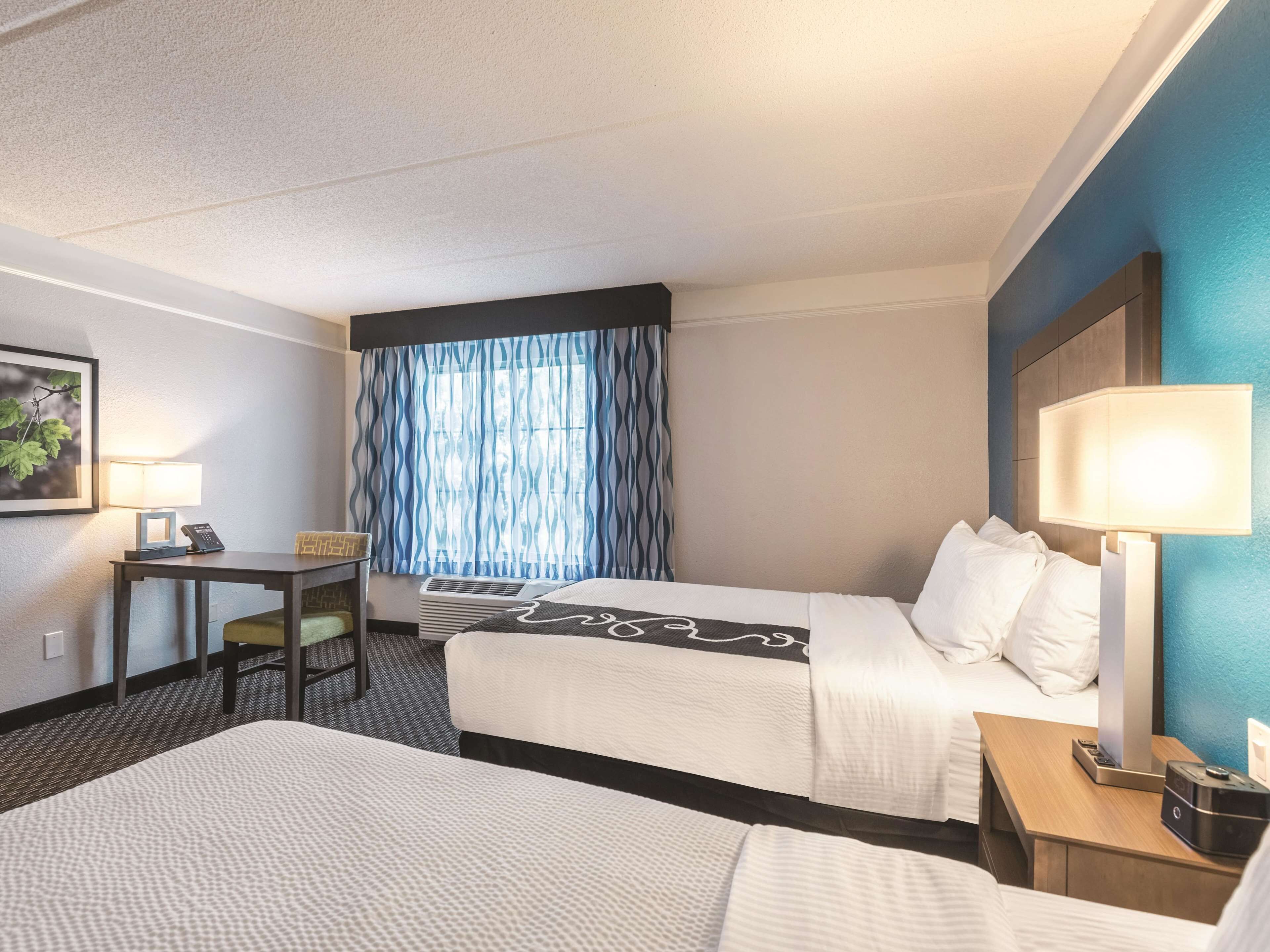 A hotel room with two beds, a desk, a chair, a lamp, and a window with curtains.