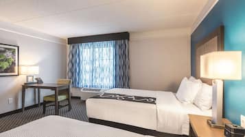 A hotel room with two beds, a desk, a chair, a lamp, and a window with curtains.