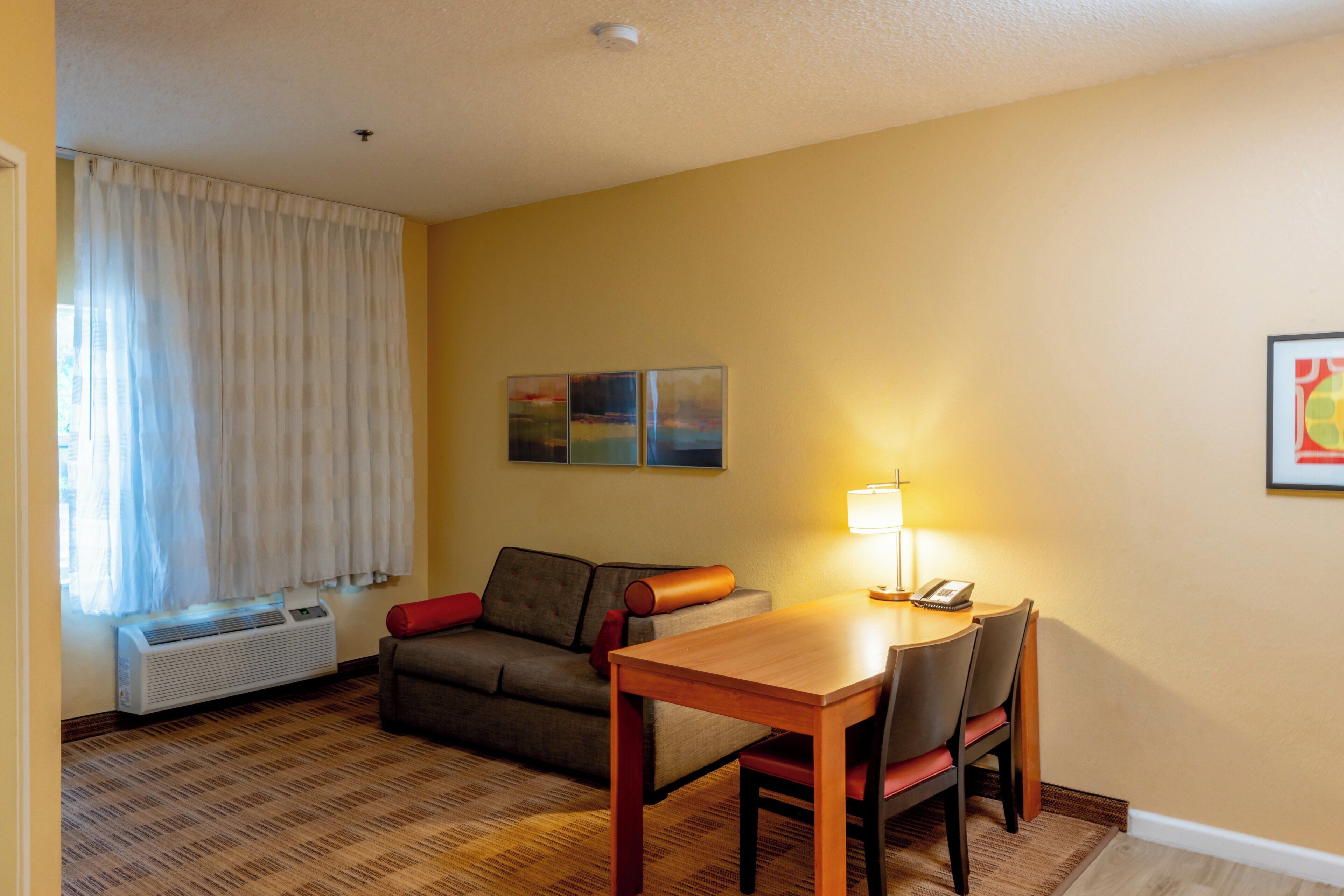 Ein Hotelzimmer mit einer Couch, einem Esstisch, Stühlen, einer Lampe, einem Telefon und einem Bild an der Wand.