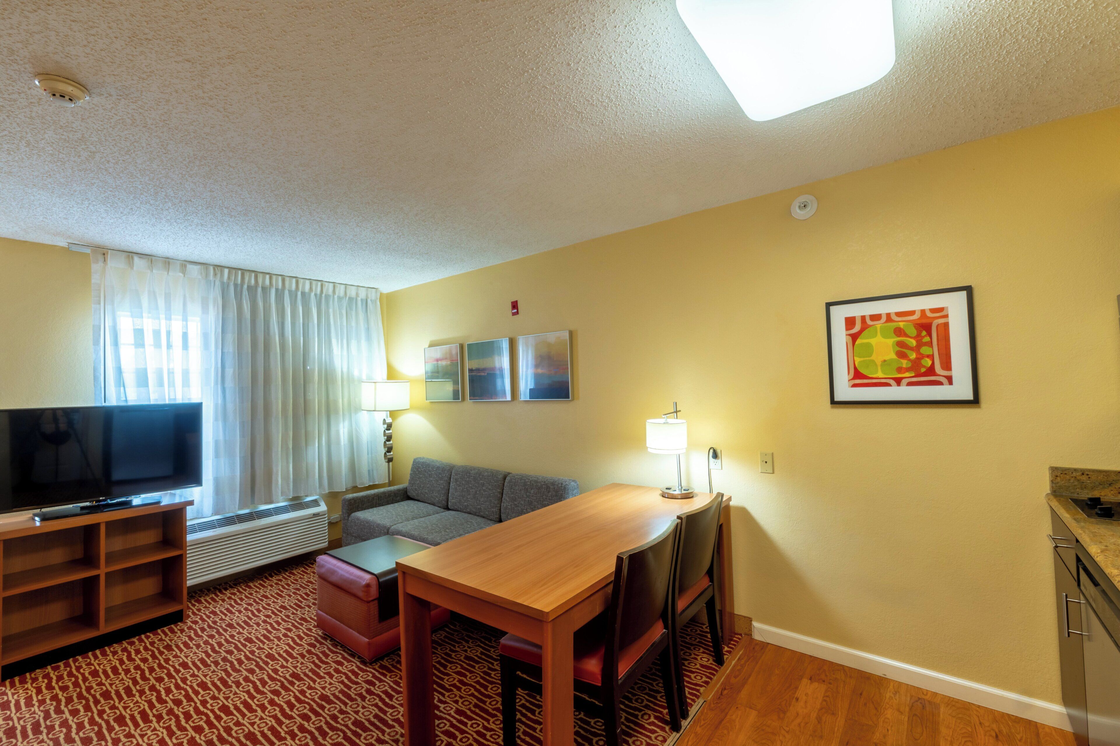 Habitación de hotel con televisor de pantalla plana, sofá, mesa de comedor, lámpara y un cuadro en la pared.