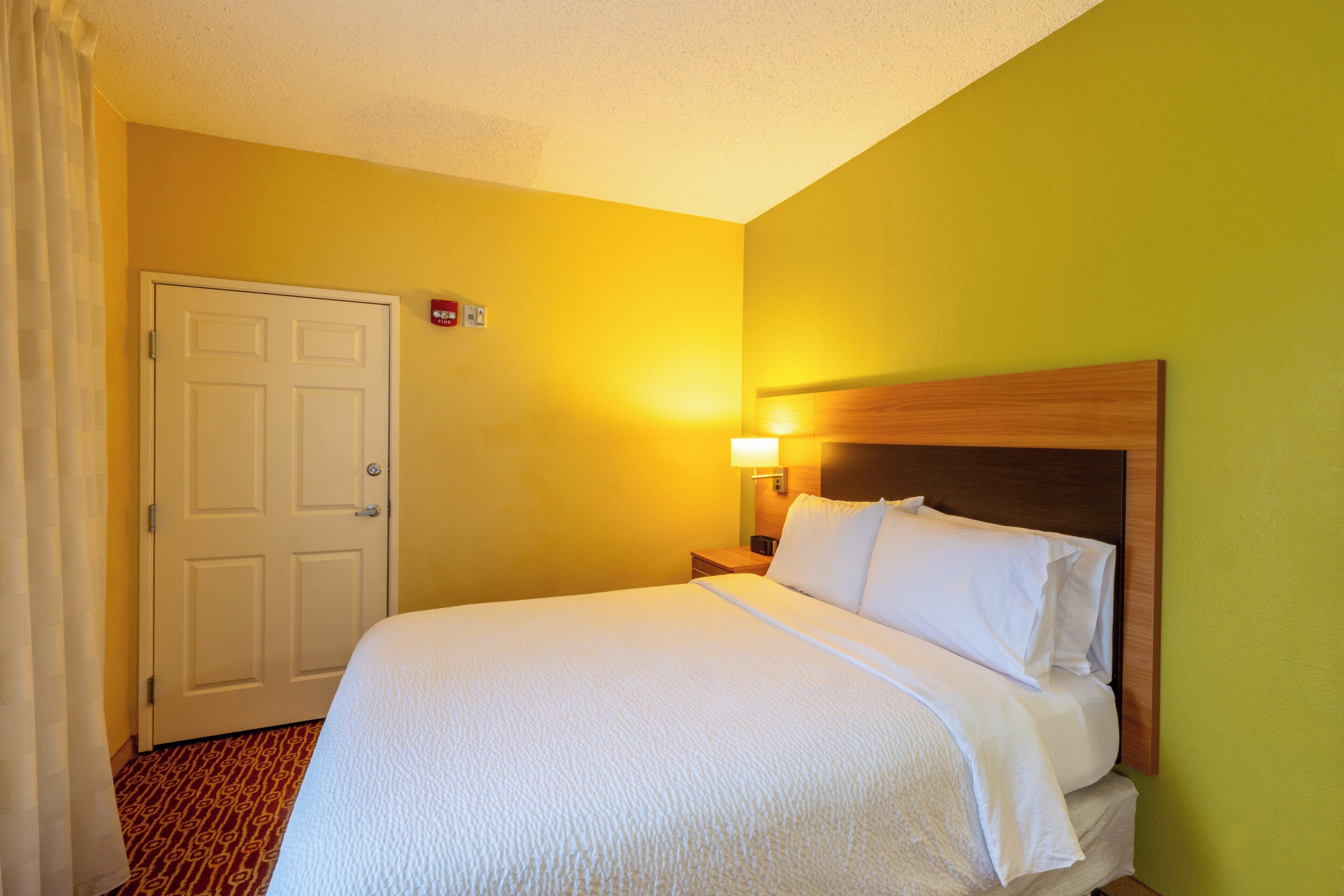 A hotel room with a bed, a nightstand, a lamp, and a door.